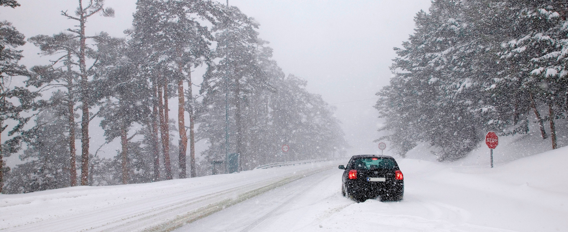 Coche negro detenido en medio de una nevada, con las luces de freno encendidas, en una carretera nevada rodeada de árboles muy altos al fondo Coche negro detenido en medio de una nevada, con las luces de freno encendidas, en una carretera nevada rodeada de árboles muy altos al fondo