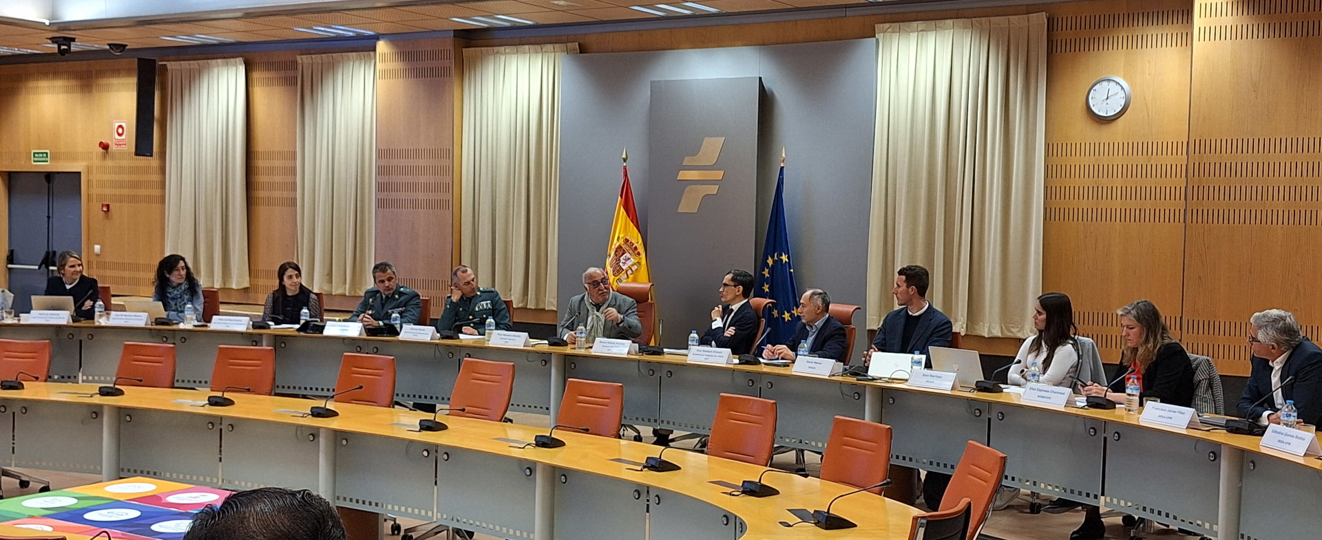 Una reunión de trabajo en una sala de conferencias semicircular con representantes institucionales y de seguridad sentados a una gran mesa. Al fondo, destacan las banderas de España y la Unión Europea junto a un logotipo institucional. Los asistentes tienen placas de identificación y micrófonos frente a ellos en un entorno profesional y formal.
