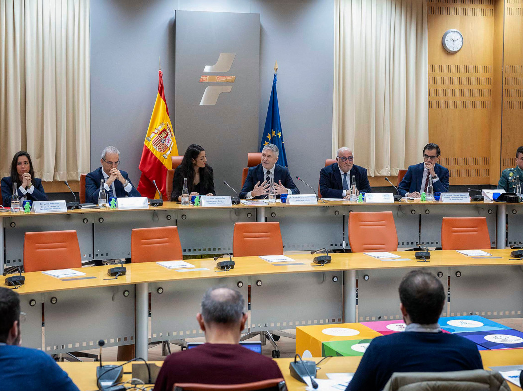 Reunión institucional con varias personas sentadas en una mesa larga; una persona habla en el centro. Al fondo se ven las banderas de España y de la Unión Europea.