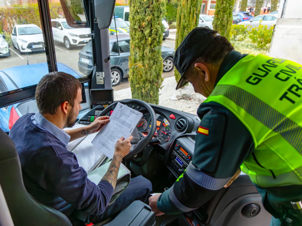 Un conductor de autobús sentado en el asiento del conductor con un papel entre sus manos y un guardia civil de pie a su lado en el interior de un autobús