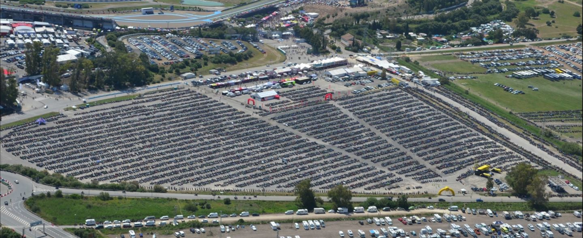 Imagen aérea de la explanada de una carretera con un parking donde se ven motocicletas aparcadas ordenadamente y unas carpas de eventos. También hay otros vehículos aparcados y vegetación.