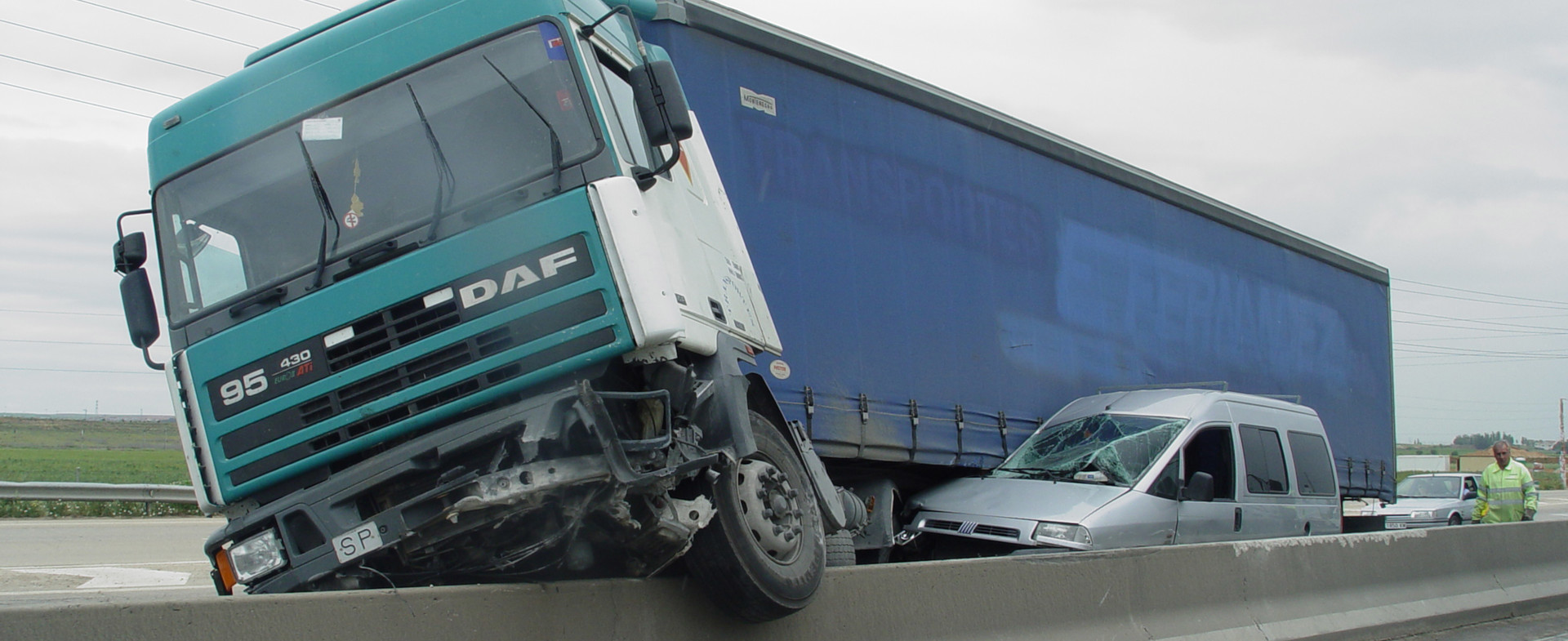 Camión accidentado sobre la barrera de una carretera, aplastando una furgoneta gris.