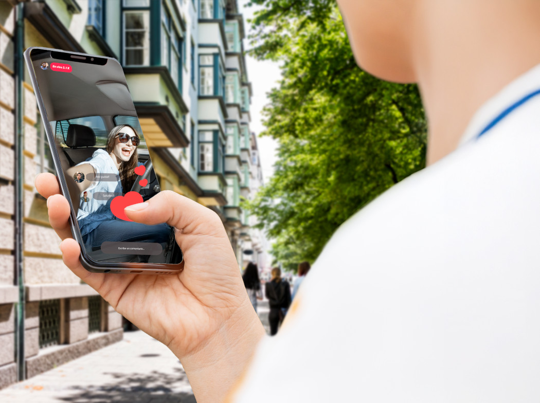 Joven con un teléfono móvil en la mano y el pulgar encima de un corazón en la pantalla de su móvil en el que hay una chica conduciendo sin cinturón de seguridad